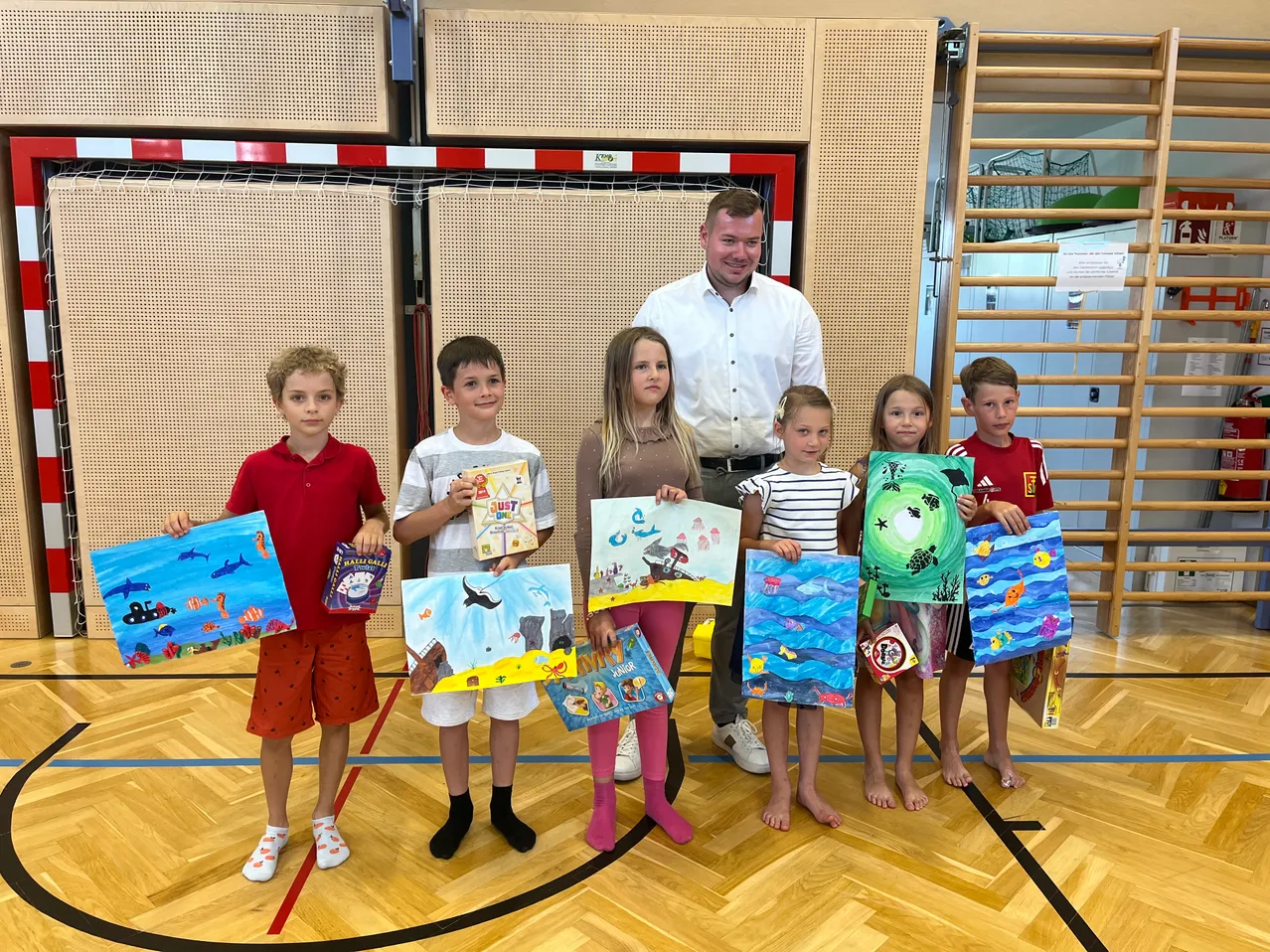 Eine Gruppe von Kindern in einer Schulsporthalle hält Kunstprojekte und ein Buch in der Hand, während ein Mann lächelnd hinter ihnen steht.