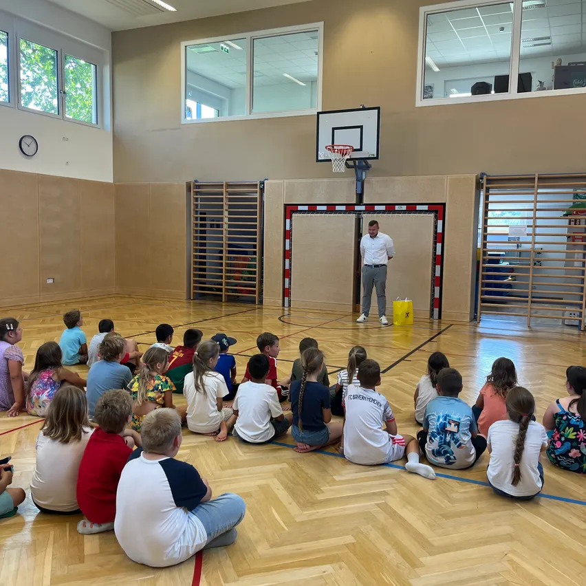 Eine Gruppe von Kindern sitzt auf dem Boden einer Basketballhalle und hört einem Mann zu, der vor dem Korb steht.