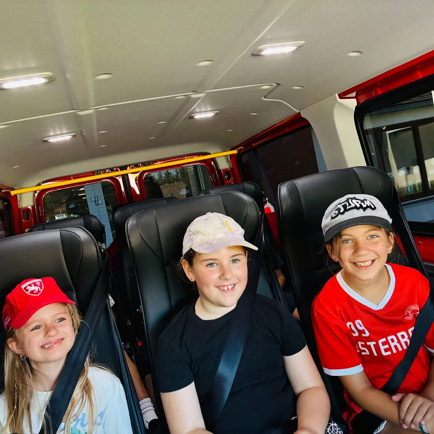 Drei Mädchen sitzen in einem roten Bus. Das Mädchen auf der linken Seite trägt eine rote Mütze. Das Mädchen in der Mitte trägt eine weiße Mütze. Das Mädchen auf der rechten Seite trägt ein rotes Trikot mit der Nummer 39.