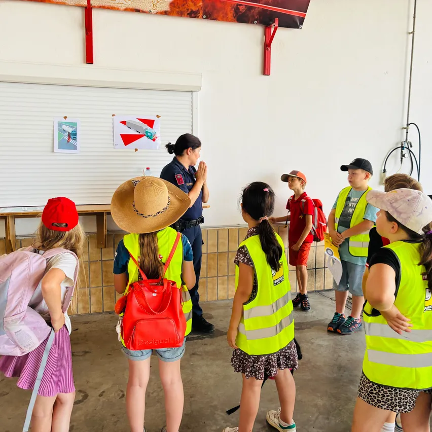 Eine Gruppe von Kindern in Sicherheitswesten und Hüten versammelt sich in einem Raum und hört einer Frau in Uniform zu. Der Raum hat weiße Wände und eine weiße Rolltür.