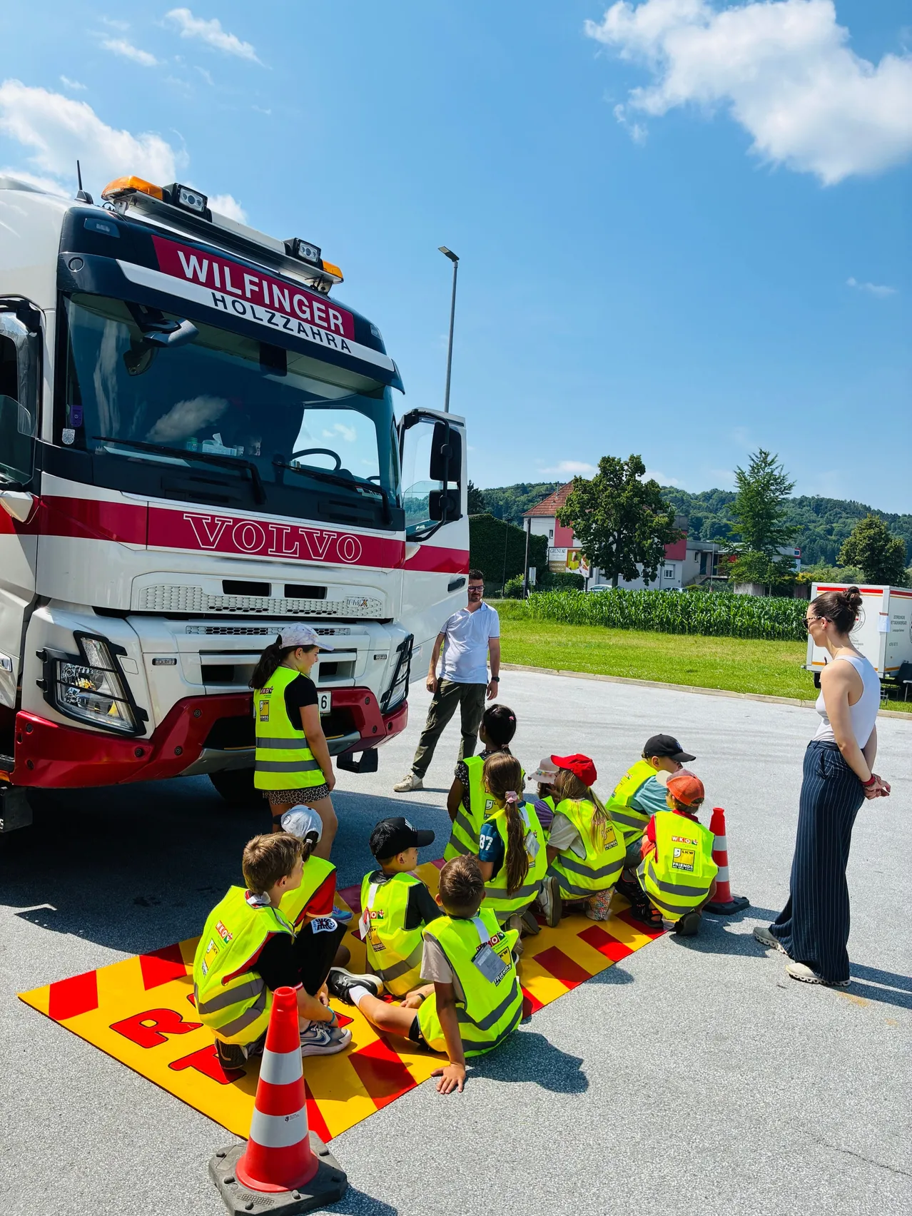 Eine Gruppe von Kindern in gelben Westen sitzt auf einer Straße und hört einem Erwachsenen zu. Ein Volvo-LKW steht in der Nähe, neben ihm steht ein Mann.