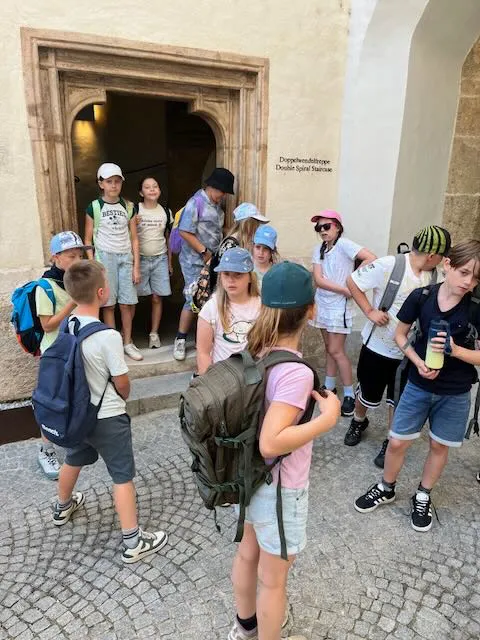 Eine Gruppe von Kindern mit Rucksäcken steht vor einem Gebäude, möglicherweise auf einer Schulfahrt. Einige tragen Mützen, andere Sonnenbrillen. Einer hält ein Getränk.