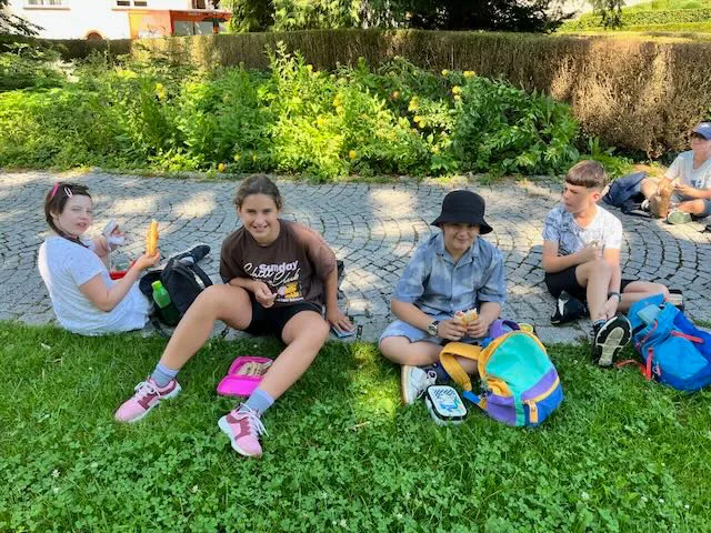 Fünf Kinder sitzen auf einem grünen Rasen in einem Außenpark, lächeln und genießen ihr Essen. Jedes Kind hat einen Rucksack neben sich. Ein Kind hat ein braunes Shirt mit weißem Text.