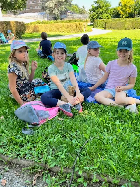 Vier Mädchen sitzen mit ihren Rucksäcken und Mützen auf der Wiese. Sie scheinen ein Picknick zu machen. Hinter ihnen sitzen weitere Menschen auf der Wiese.