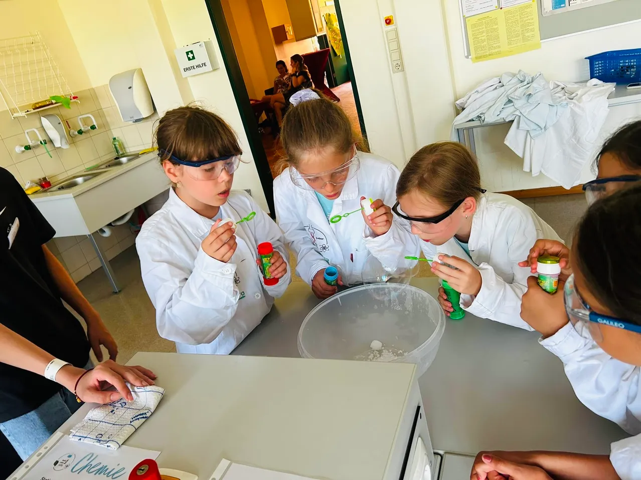 Vier Kinder in Laborkitteln und Schutzbrillen führen ein Experiment in einem Labor durch. Sie halten grüne Flaschen und Röhrchen und mischen etwas in einer Schüssel auf einem Tisch. Dahinter befindet sich ein Waschbecken und ein Durchgang zu einem Raum.