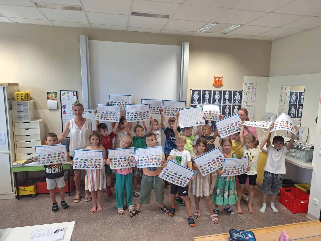 Eine Gruppe von Kindern und zwei Frauen in einem Klassenzimmer halten bunte Papiere hoch. Die Frau in Weiß steht vorne.