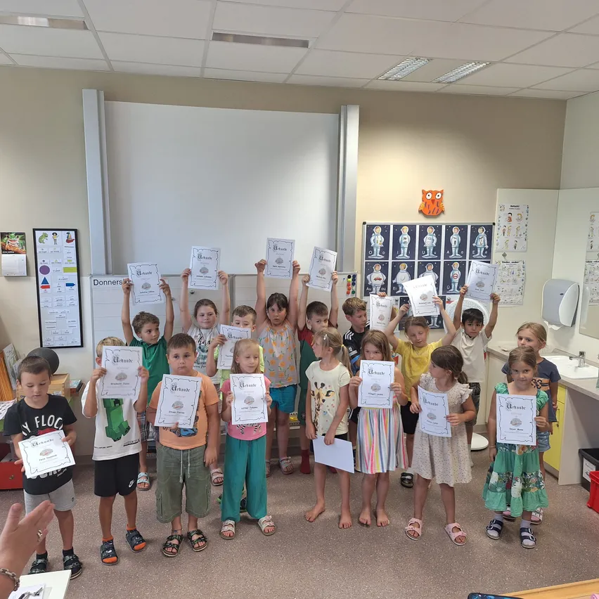 Ein Klassenzimmer voller Kinder, die Urkunden halten und lächeln, mit einer Tafel, Dekorationen und einem Waschbecken im Hintergrund.