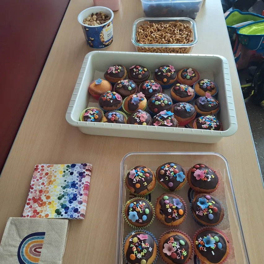 Ein hölzerner Tisch hat ein Tablett mit Schokoladen-Cupcakes, zwei Behälter mit Snacks, eine Schüssel Müsli und zwei bunte Servietten.