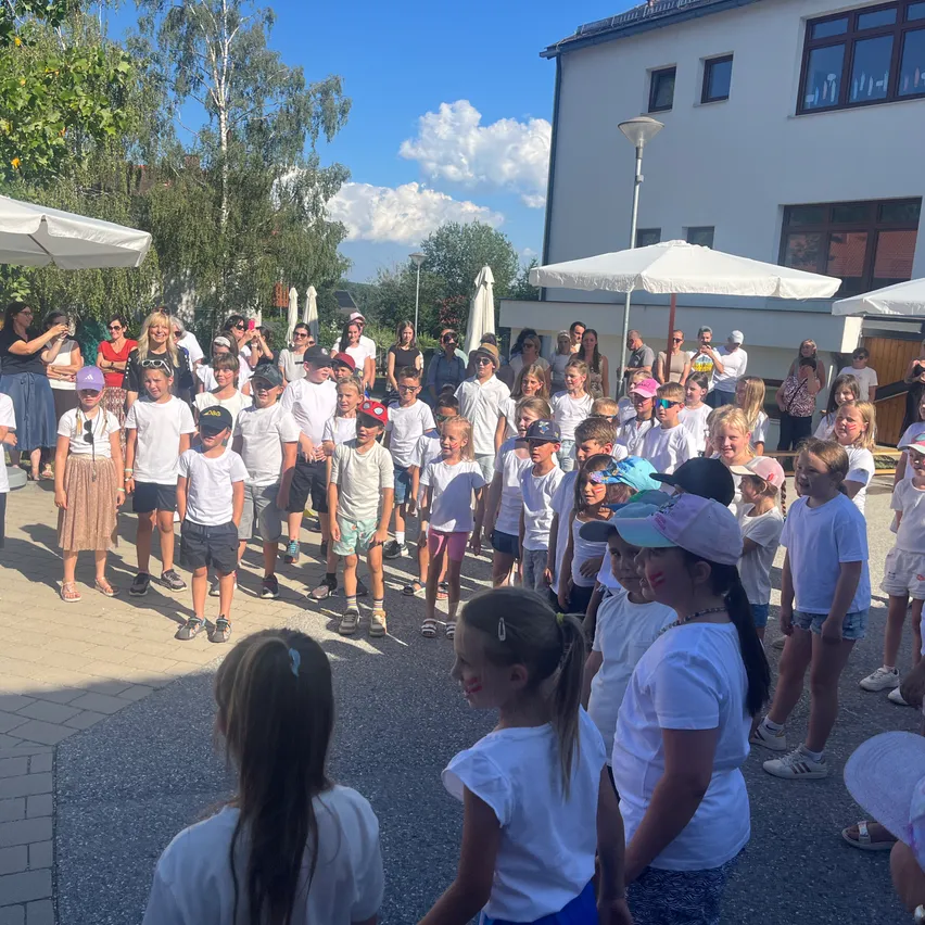 Eine Gruppe von Kindern und Erwachsenen in weißen T-Shirts versammelt sich draußen in der Nähe eines Gebäudes, einige tragen Mützen und Sonnenbrillen.