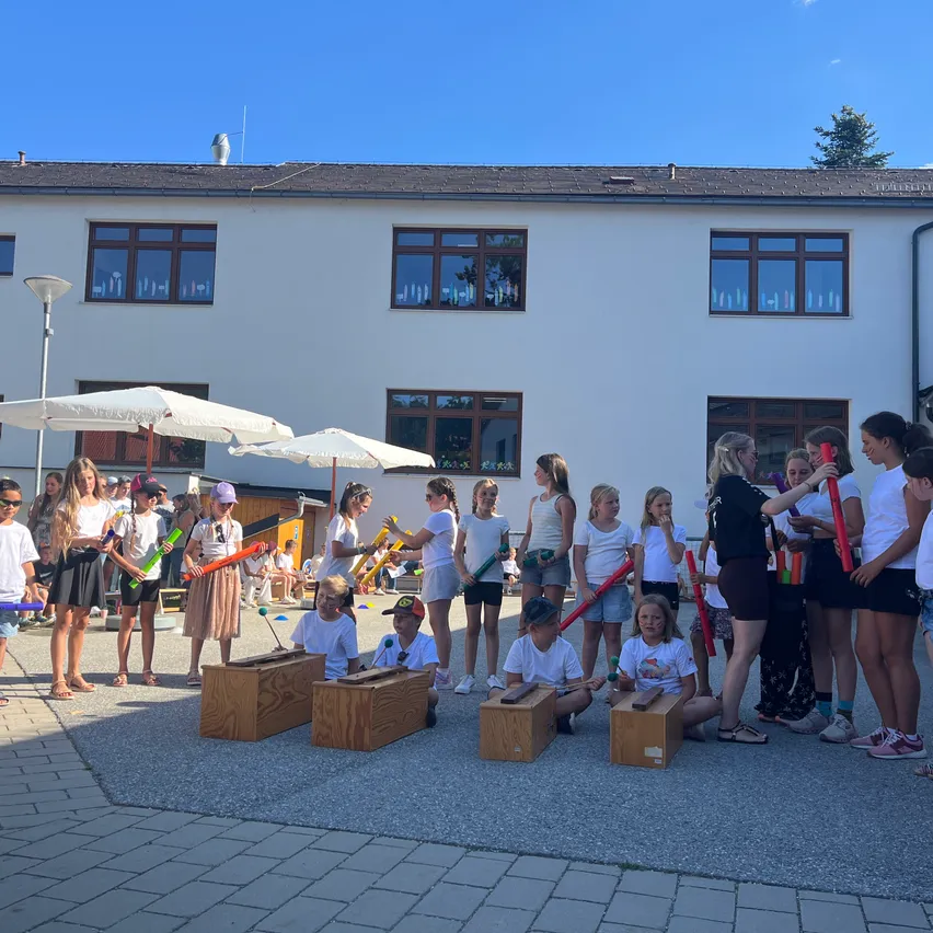 Eine Gruppe von Kindern ist draußen versammelt, jedes hält ein Musikinstrument. Sie sind vor Holzkisten angeordnet und scheinen eine Aufführung zu machen. Die Szene spielt sich vor einem weißen Gebäude mit mehreren Fenstern ab.