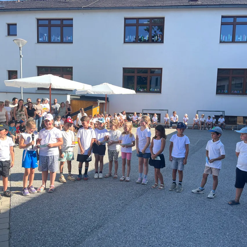 Eine Gruppe von Kindern steht in einer Reihe auf einem Kiesweg, trägt Hüte und Turnschuhe. Dahinter hält ein Erwachsener ein Mikrofon. Ein Gebäude mit Glasfenstern ist im Hintergrund.