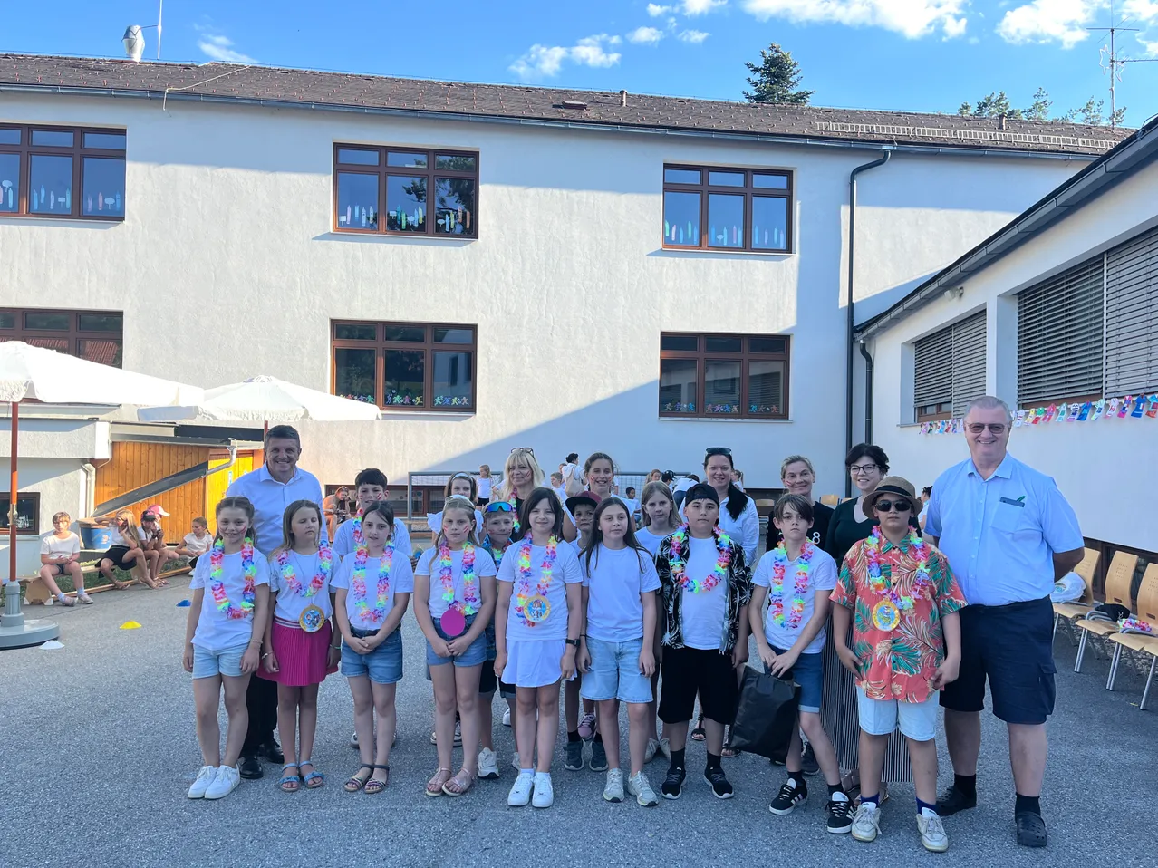 Eine Gruppe von Kindern in passenden T-Shirts und Blumenkränzen posiert für ein Foto vor einem Gebäude. Zwei Männer und zwei Frauen stehen an den Seiten.