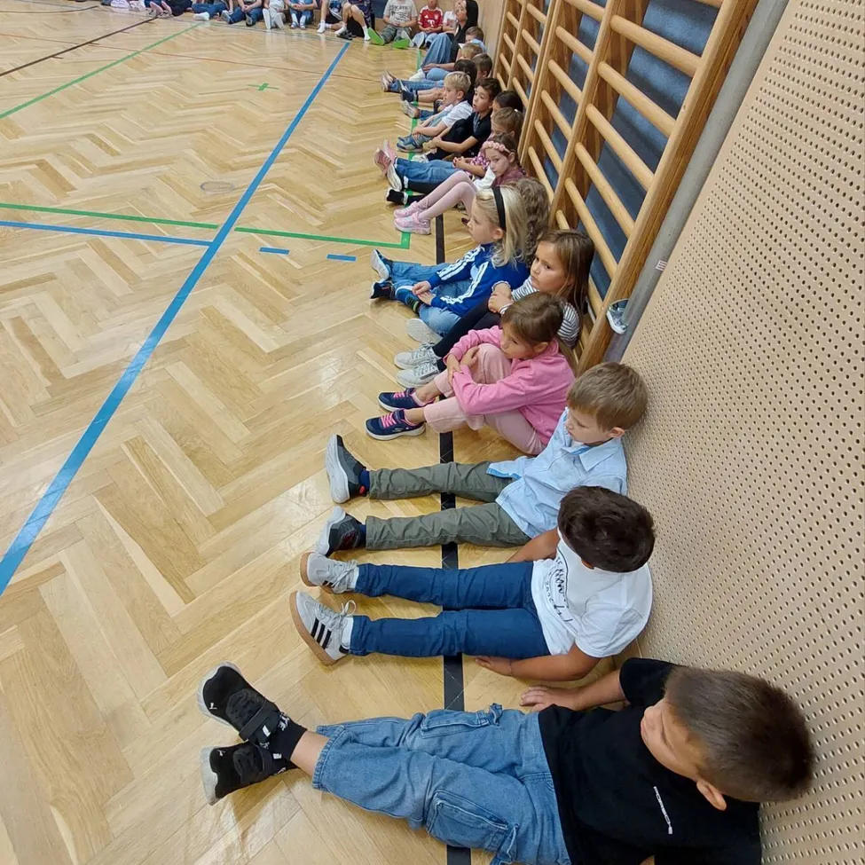 Eine Gruppe von Kindern sitzt in einer Reihe auf dem Boden einer Turnhalle, einige lehnen sich gegen die Wand.
