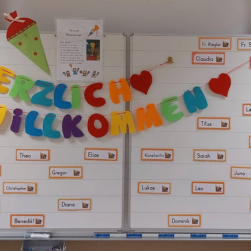 Eine weiße Tafel mit einem bunten Banner, das 'Herzlich Willkommen' mit Herzen und Namen von Schülern darauf befestigt ist.