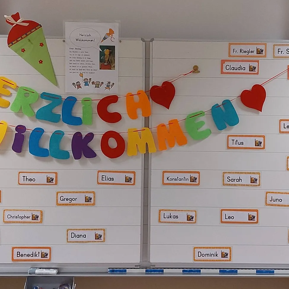 Eine weiße Tafel mit einem bunten Banner, das 'Herzlich Willkommen' mit Herzen und Namen von Schülern darauf befestigt ist.
