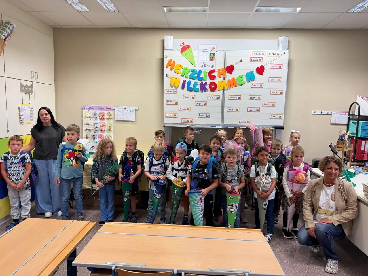 Eine Gruppe von Kindern und zwei Erwachsenen stehen in einem Klassenzimmer. Die Kinder tragen Rucksäcke und halten Banner. Eine Frau steht hinter ihnen und eine andere Frau sitzt auf dem Boden.
