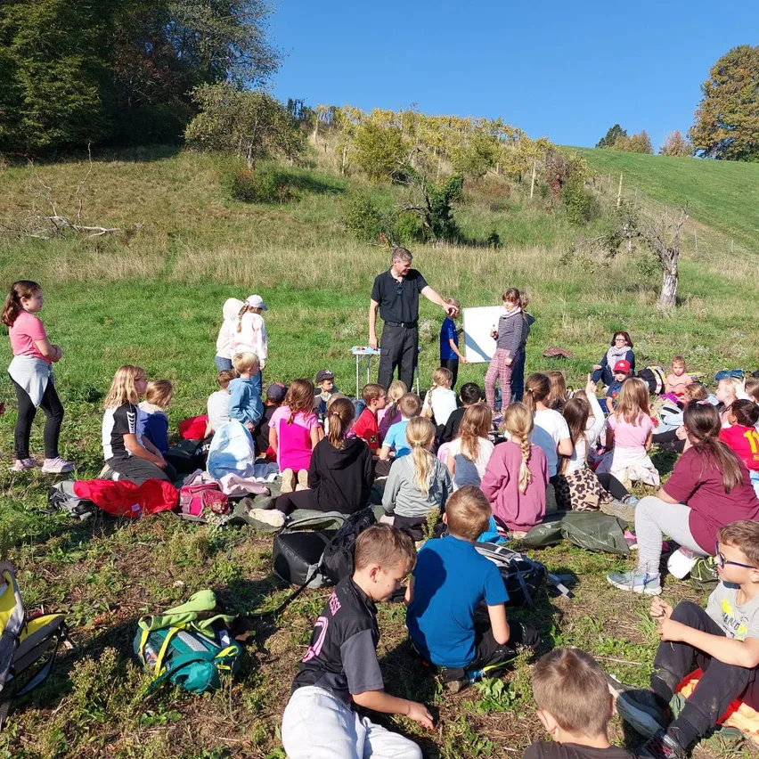 Eine Gruppe von Kindern und Erwachsenen sitzt auf einem Grashügel unter einem klaren blauen Himmel. Ein Mann steht vor ihnen und hält ein Whiteboard.