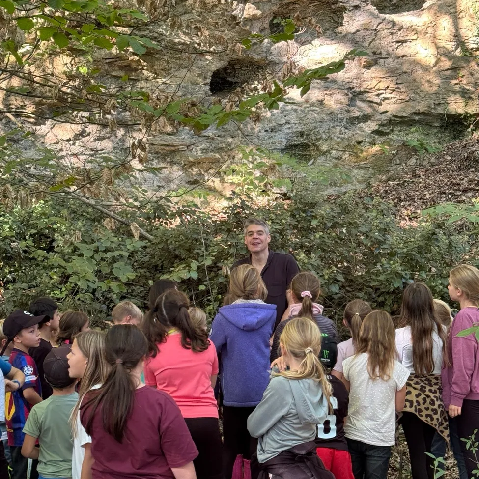 Eine Gruppe von Kindern und ein erwachsener Mann stehen vor einer Felsformation in einem Wald.
