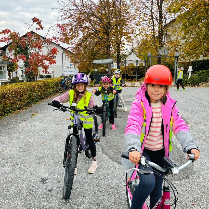 Eine Gruppe von Kindern fährt auf einem gepflasterten Weg mit Fahrrädern. Sie tragen alle Helme und reflektierende Westen. Die Kinder lächeln und scheinen ihre Fahrt zu genießen.