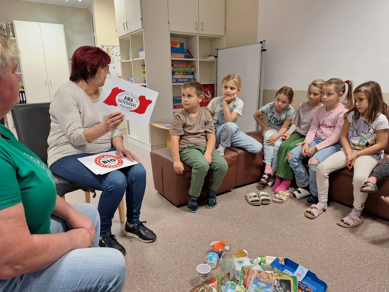 Eine Gruppe von Kindern sitzt auf einer Couch und hört einer Frau zu, die ein Plakat hält.