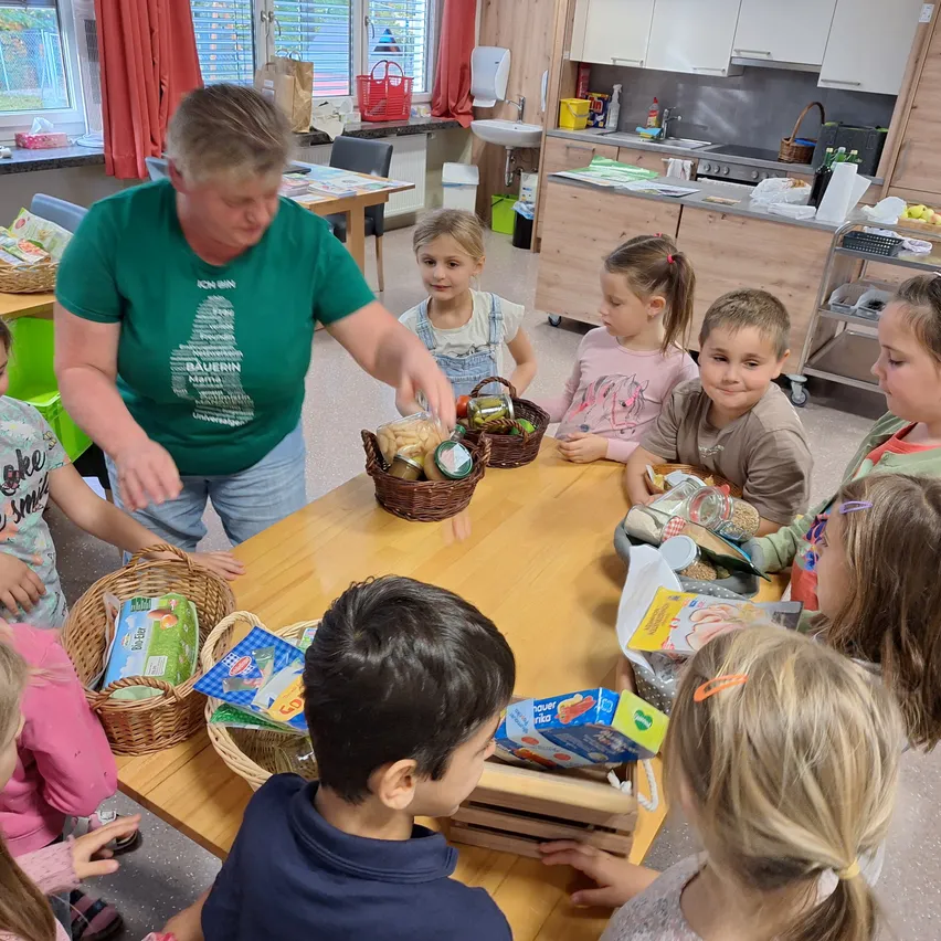 Eine Frau in einem grünen T-Shirt steht vor einem Tisch mit einer Gruppe von Kindern. Sie halten alle Körbe voller Essen.