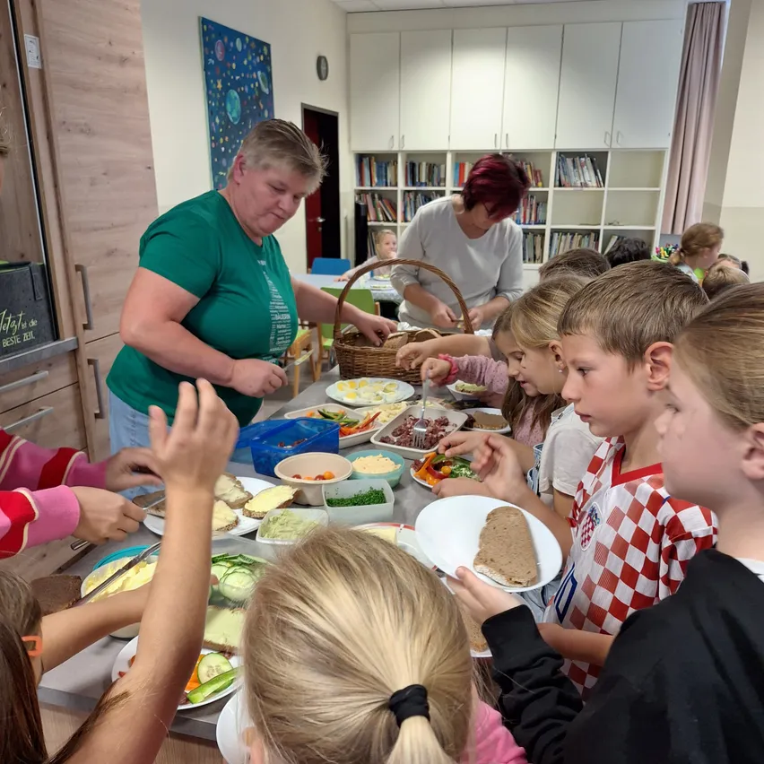 Eine Gruppe von Kindern und zwei Frauen versammelt sich um einen Tisch voller verschiedener Speisen. Eine Frau serviert Essen in Schüsseln, während die andere Frau hinter ihr steht.