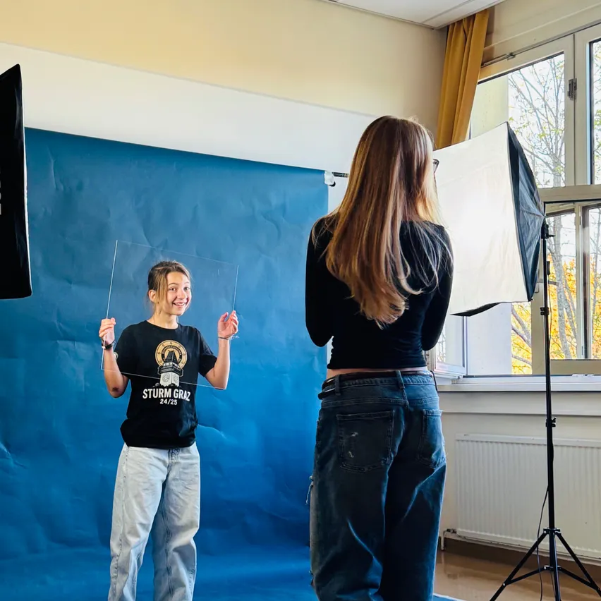 Ein junges Mädchen steht vor einem blauen Hintergrund in einem Studio und hält ein durchsichtiges Plastikblatt. Eine Frau mit langen Haaren steht hinter ihr und schaut sie an.