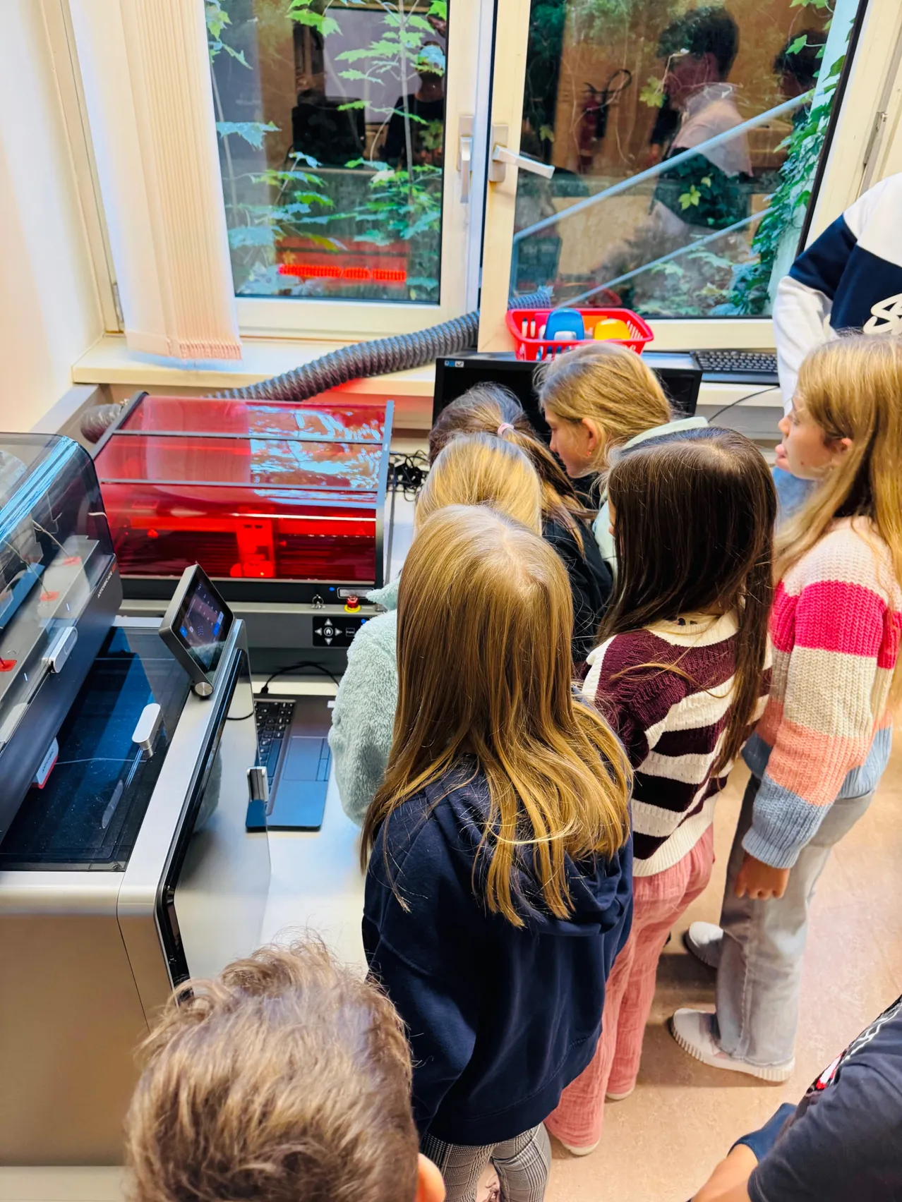 Eine Gruppe von Kindern steht um einen großen roten 3D-Drucker in einem Klassenzimmer.