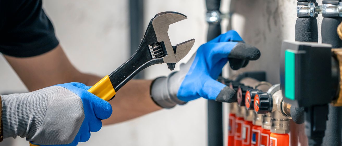 Eine Person mit blauen Handschuhen hält eine Schraubenschlüssel in der Nähe eines Feuerlöschers. Der Hintergrund ist unscharf, was auf einen Fokus auf die Hände und Werkzeuge hinweist.