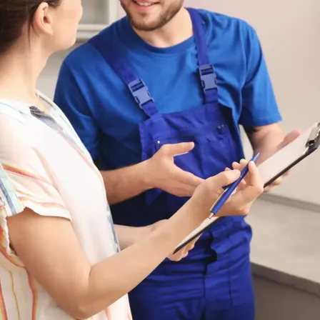 Eine Frau und ein Mann in blauen Uniformen sind in einer Küche. Der Mann spricht mit der Frau, während sie auf eine Clipboard schreibt.