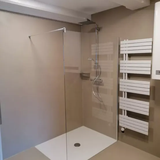 Ein modernes Badezimmer verfügt über eine Glasdusche mit silberner Duschbrause, ein eingebautes Regal und einen weißen Handtuchhalter. Die Wände sind hellbeige und der Boden weiß.