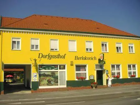Ein gelbes Gebäude mit rotem Rand und rotem Dach namens Dorfgasthof Berlakovich. Es gibt Fenster und eine Tür mit einem Schild. Es gibt Blumen in den Fensterbänken.
