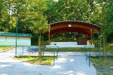 Eine Freiluftbühne mit einem geschwungenen Dach befindet sich in einem Park, umgeben von Bäumen und Grün. Links ist ein kleines Gebäude mit einem grünen Dach zu sehen.