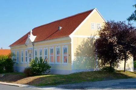 Ein gelbes Haus mit rotem Dach und weißer Einfassung steht auf einem Hügel, umgeben von Grün und einem klaren blauen Himmel.