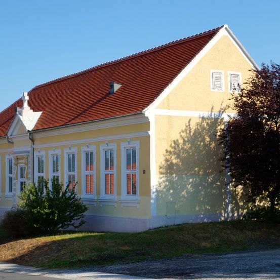 Ein gelbes Gebäude mit einem roten Dach und mehreren Fenstern, umgeben von Gras und einem Baum. Das Gebäude hat einen klassischen Architekturstil.