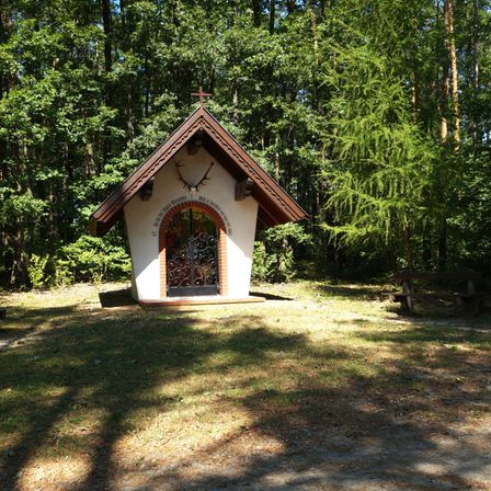 Eine kleine Holzkapelle mit einem Kreuz auf dem Dach befindet sich in einem Wald. Sie hat eine Glastür und einen Hirschkopf an der Wand befestigt.