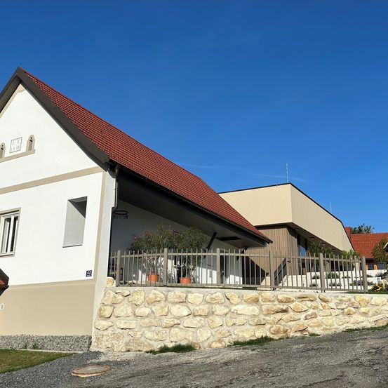 Ein Haus mit einem roten Dach und weißen Wänden. Ein Steinwall umgibt das Haus, und davor steht ein Zaun. Zwei Pflanzen in Töpfen befinden sich in der Nähe des Zauns. Ein Gebäude mit einem braunen Dach befindet sich rechts.