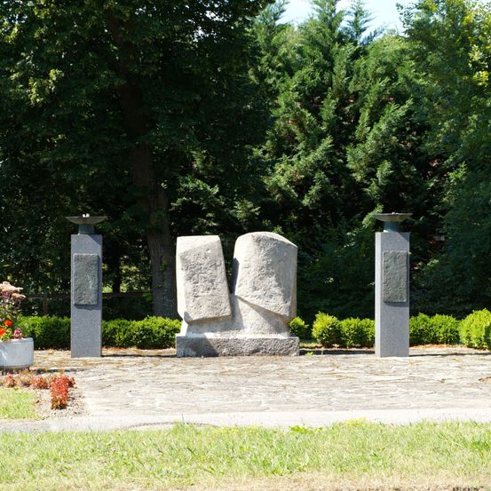 Eine Steinskulptur steht zwischen zwei Betonsockeln mit Metallschüsseln oben. Grüne Büsche umgeben den Bereich, mit Bäumen im Hintergrund.