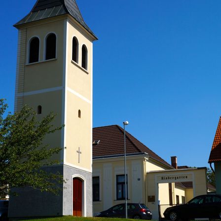 Ein hoher Kirchturm mit einem Kreuz darauf steht neben einem Gebäude. Zwei Autos sind vor dem Gebäude geparkt. Eine Straßenlaterne steht vor dem Gebäude.
