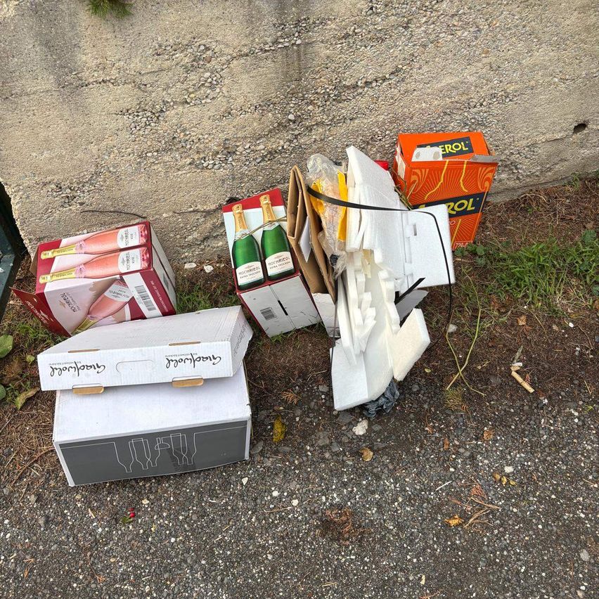 Verschiedene leere Weinkartons liegen auf dem Boden in der Nähe einer Betonmauer. Ein Kasten enthält zwei Champagnerflaschen. Ein weiterer Kasten hat einen zerbrochenen Deckel und eine Rolle Papiertücher.