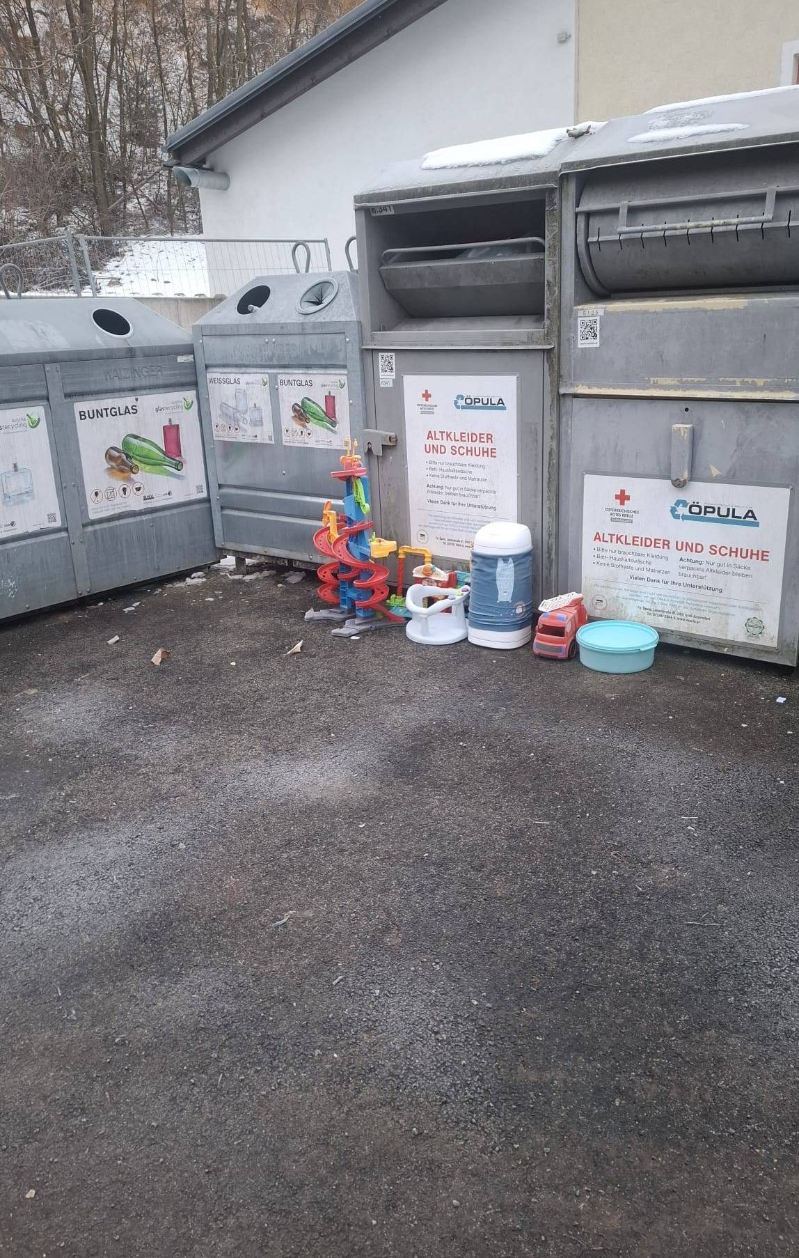 Drei Recyclingbehälter sind aufgereiht mit Schildern für Glas, Abfall und Kleidung. Ein Haufen Spielzeug, ein blauer Plastikbehälter und ein Wasserkanister liegen auf dem Boden.