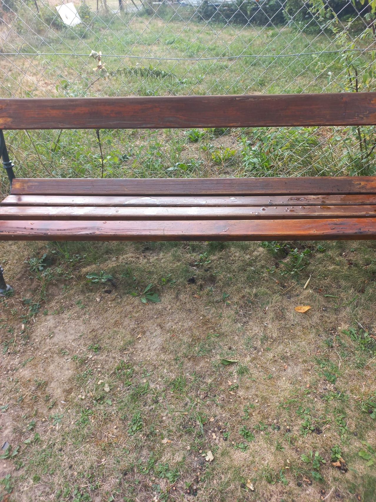 Eine alte Holzbank mit Metallbeinen steht auf einem Grasbereich in der Nähe eines Zauns. Die Bank weist deutliche Abnutzungsspuren auf.