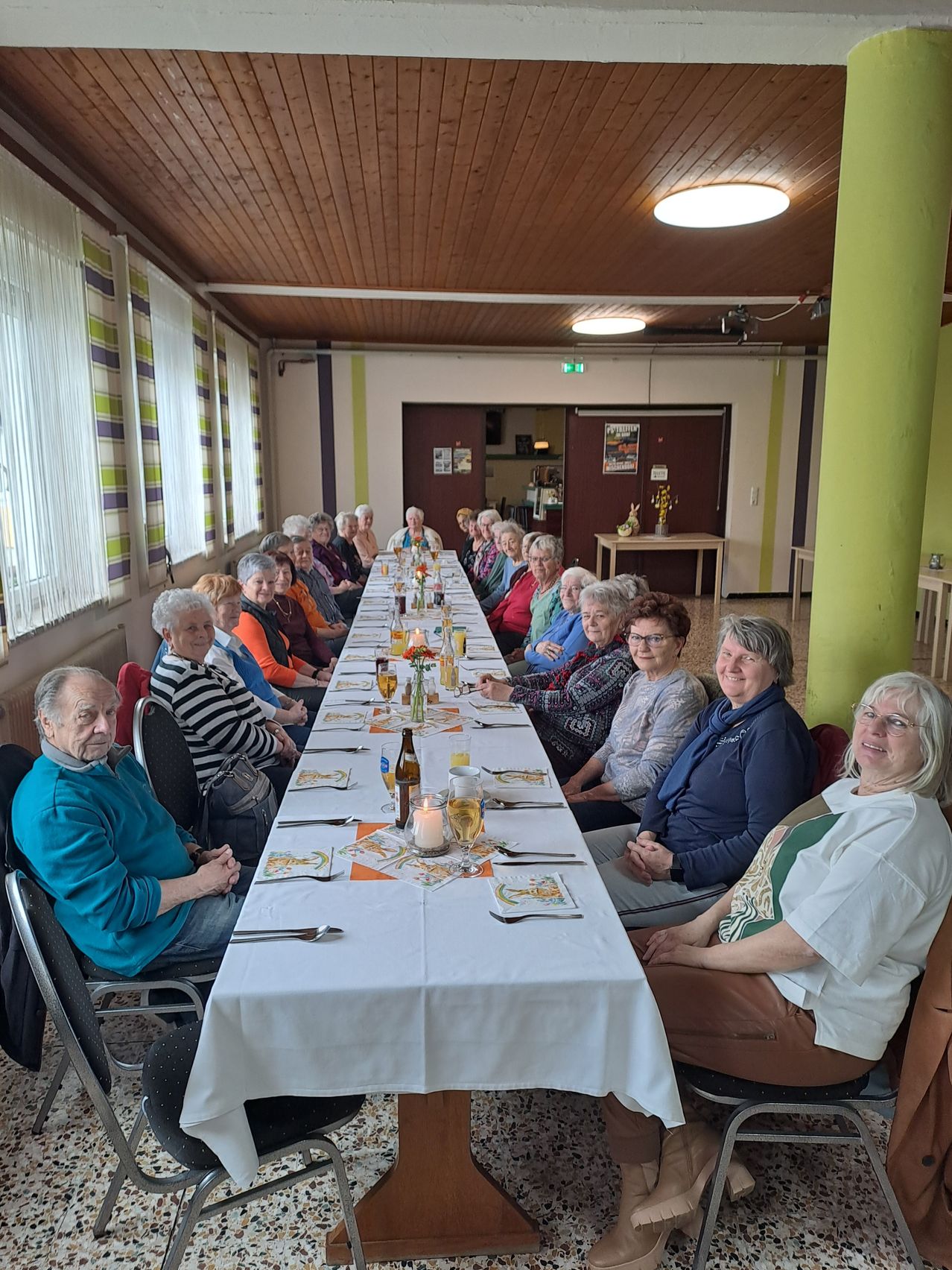Eine Gruppe älterer Frauen und Männer sitzt an einem langen Tisch und lächelt für ein Gruppenfoto.