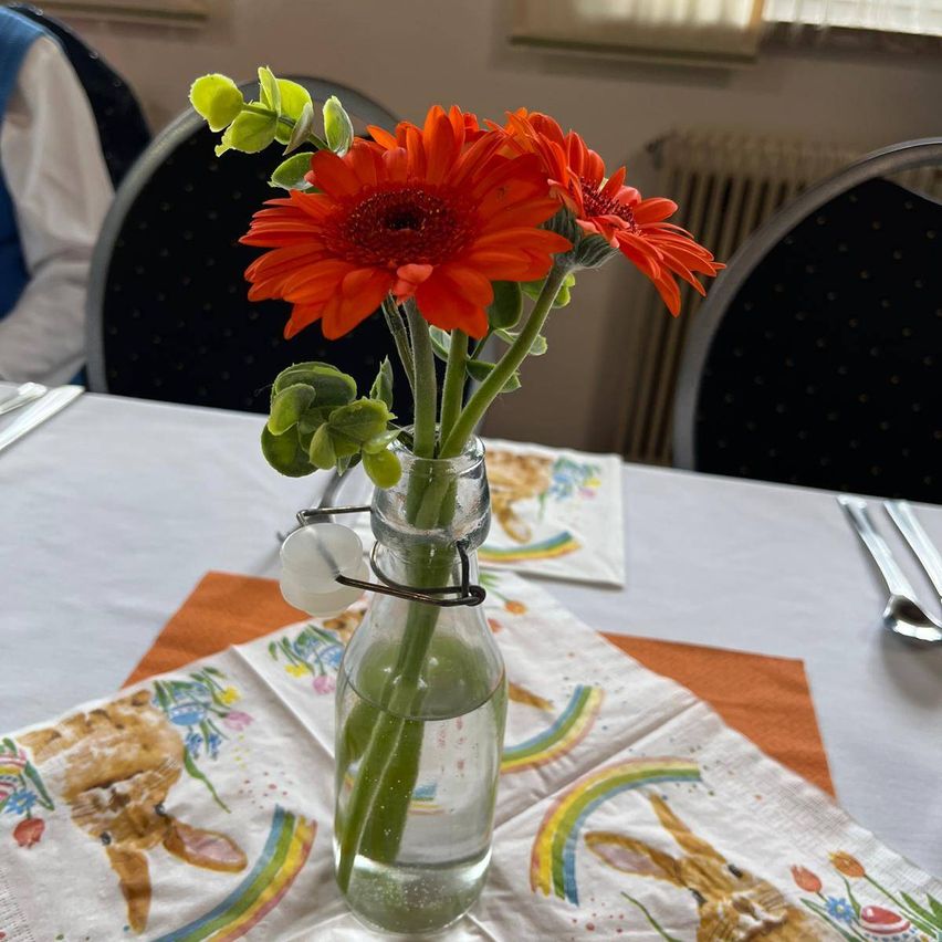 Zwei orangefarbene Gänseblümchen in einer Glasflasche mit Wasser auf einem Tisch, der mit einer Serviette mit einem Hasenbild bedeckt ist.