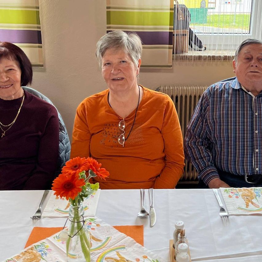 Drei Menschen sitzen lächelnd an einem Tisch. Auf dem Tisch stehen eine Blumenvase, Besteck und Gewürze. Hinter ihnen befindet sich ein Fenster mit Jalousien.