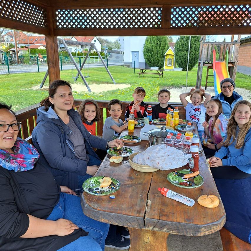 Eine Gruppe von Erwachsenen und Kindern sitzt um einen Holztisch herum, möglicherweise in einem Park, mit Essen und Getränken auf dem Tisch. Dahinter befindet sich ein Spielplatz mit einer Rutsche.