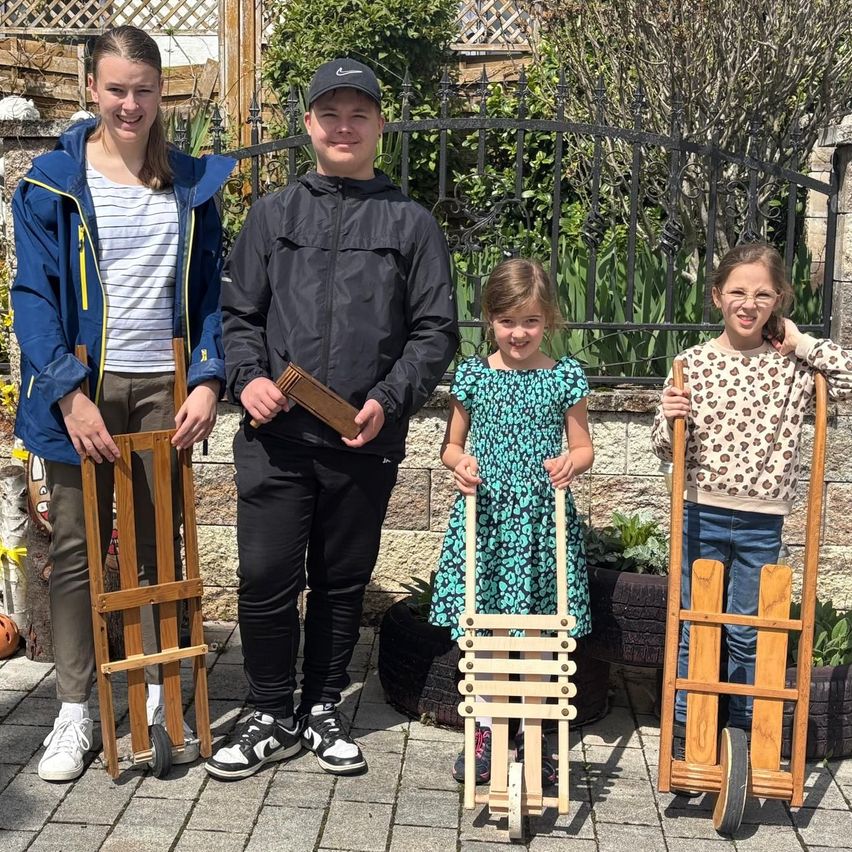 Vier Kinder, zwei Mädchen und zwei Jungen, stehen draußen in einem Garten. Sie halten alle Holzwagen. Das Mädchen rechts trägt eine Brille. Das Mädchen in der Mitte hält einen Wagen mit Rädern. Der Junge links hält ein Holzstück.