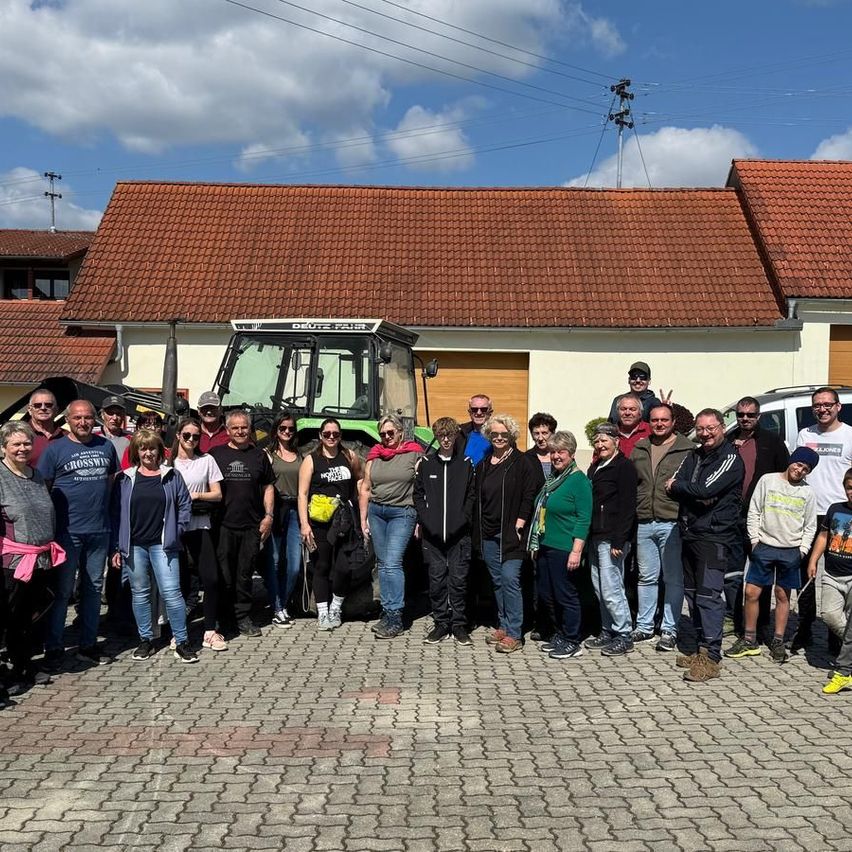 Eine große Gruppe von Menschen posiert für ein Foto vor einem Haus mit rotem Dach, einem Traktor und elektrischen Leitungen.