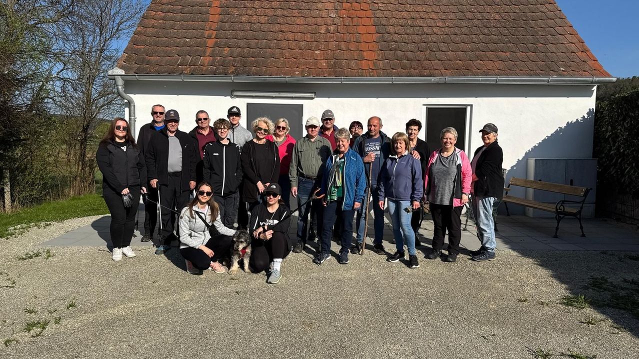 Eine Gruppe von Menschen steht und posiert für ein Foto vor einem Gebäude. Einige Leute halten Stöcke, und eine Frau hält einen Hund.