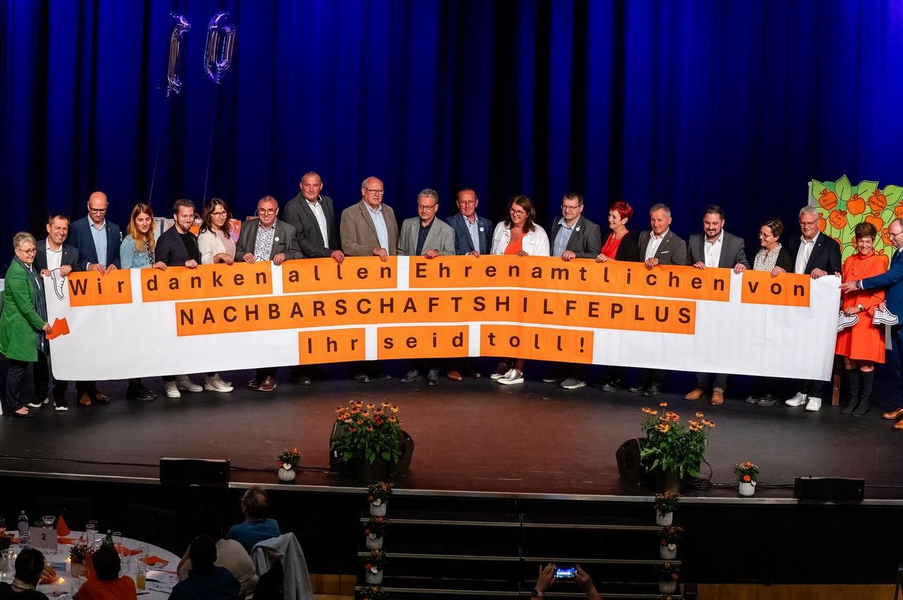 Eine Gruppe von Menschen steht auf einer Bühne und hält ein Banner. Es steht 'Danken allen Ehrenamtlichen Nachbarschaftshilfe Plus. Ihr seid toll!' in orange Buchstaben. Hinter ihnen sind Luftballons.